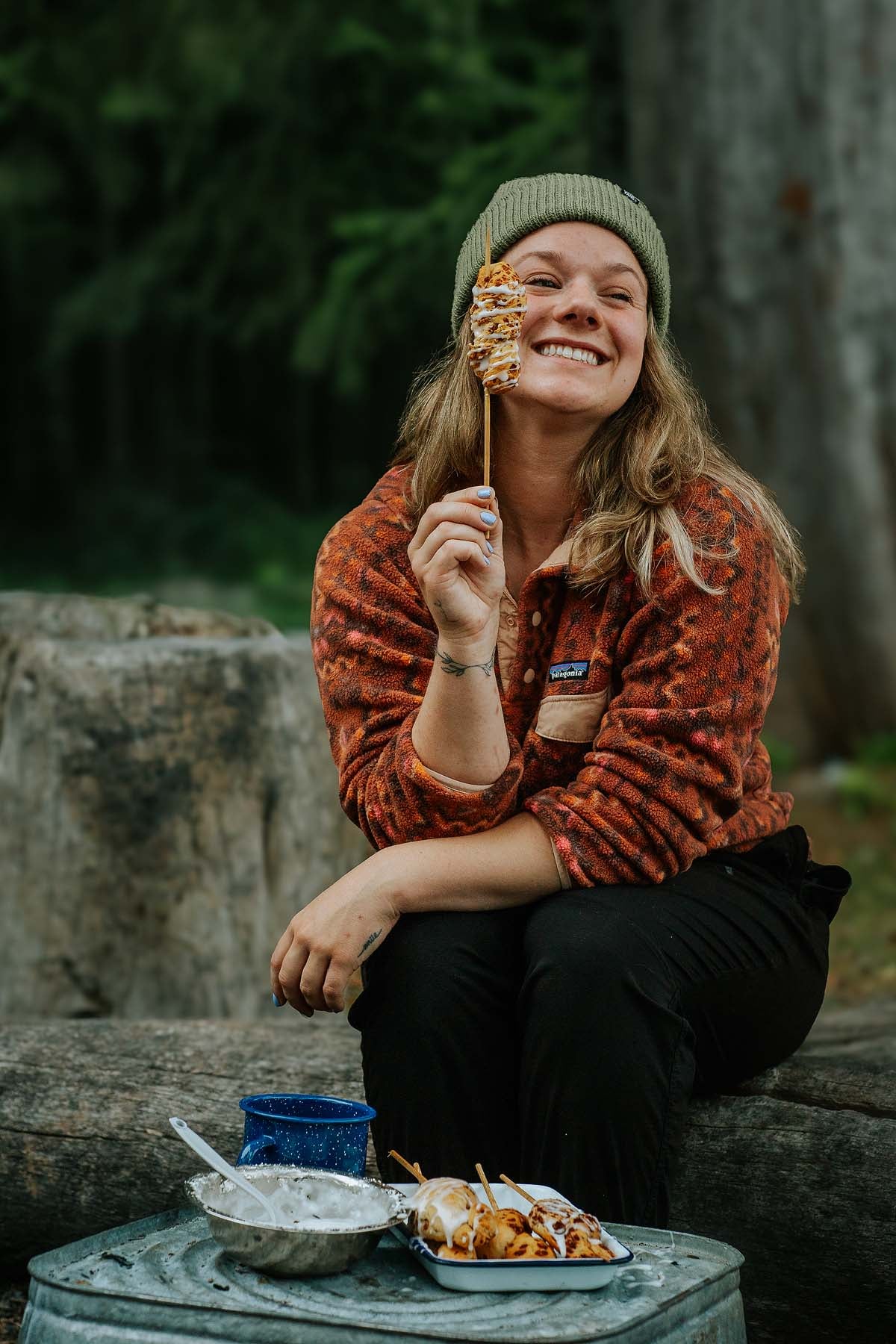 Smiling person in a beanie holds a skewer of campfire cinnamon twists while sitting outdoors at a picnic table.