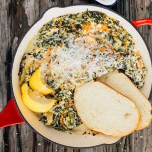 A skillet with campfire spinach artichoke dip, lemon wedges, shredded cheese, and two slices of bread on a rustic wooden table.