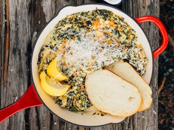 A skillet with campfire spinach artichoke dip, lemon wedges, shredded cheese, and two slices of bread on a rustic wooden table.