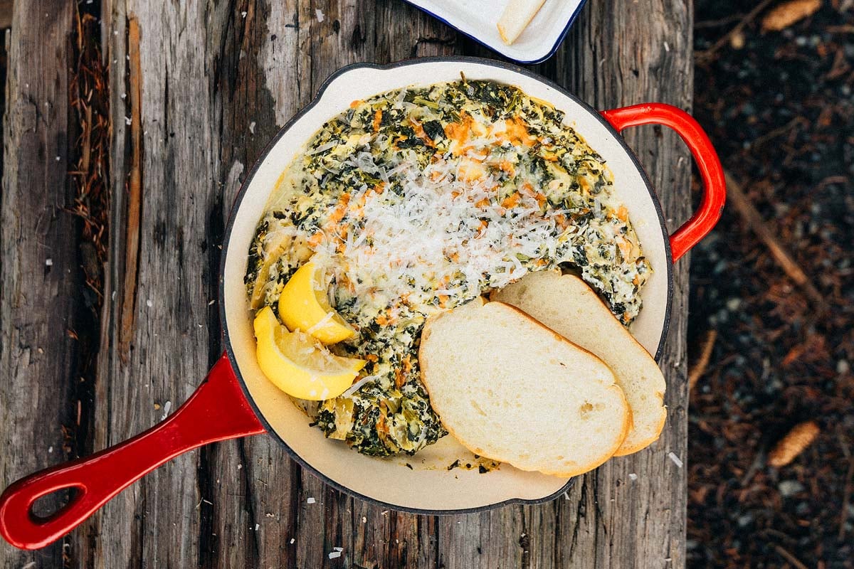 A skillet with campfire spinach artichoke dip, lemon wedges, shredded cheese, and two slices of bread on a wooden surface.