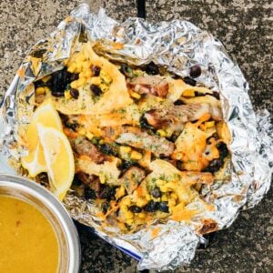 Campfire Steak and Black Bean Nachos with melted cheese, beans, and corn on foil, served with lemon wedges and a bowl of yellow soup.