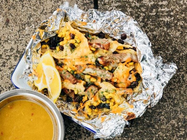 Campfire Steak and Black Bean Nachos with melted cheese, beans, and corn on foil, served with lemon wedges and a bowl of yellow soup.