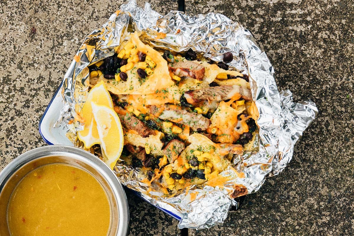 Foil packet with Campfire Steak and Black Bean Nachos&mdash;grilled meat, corn, beans, cheese, and lemon wedges&mdash;next to a bowl of yellow soup.