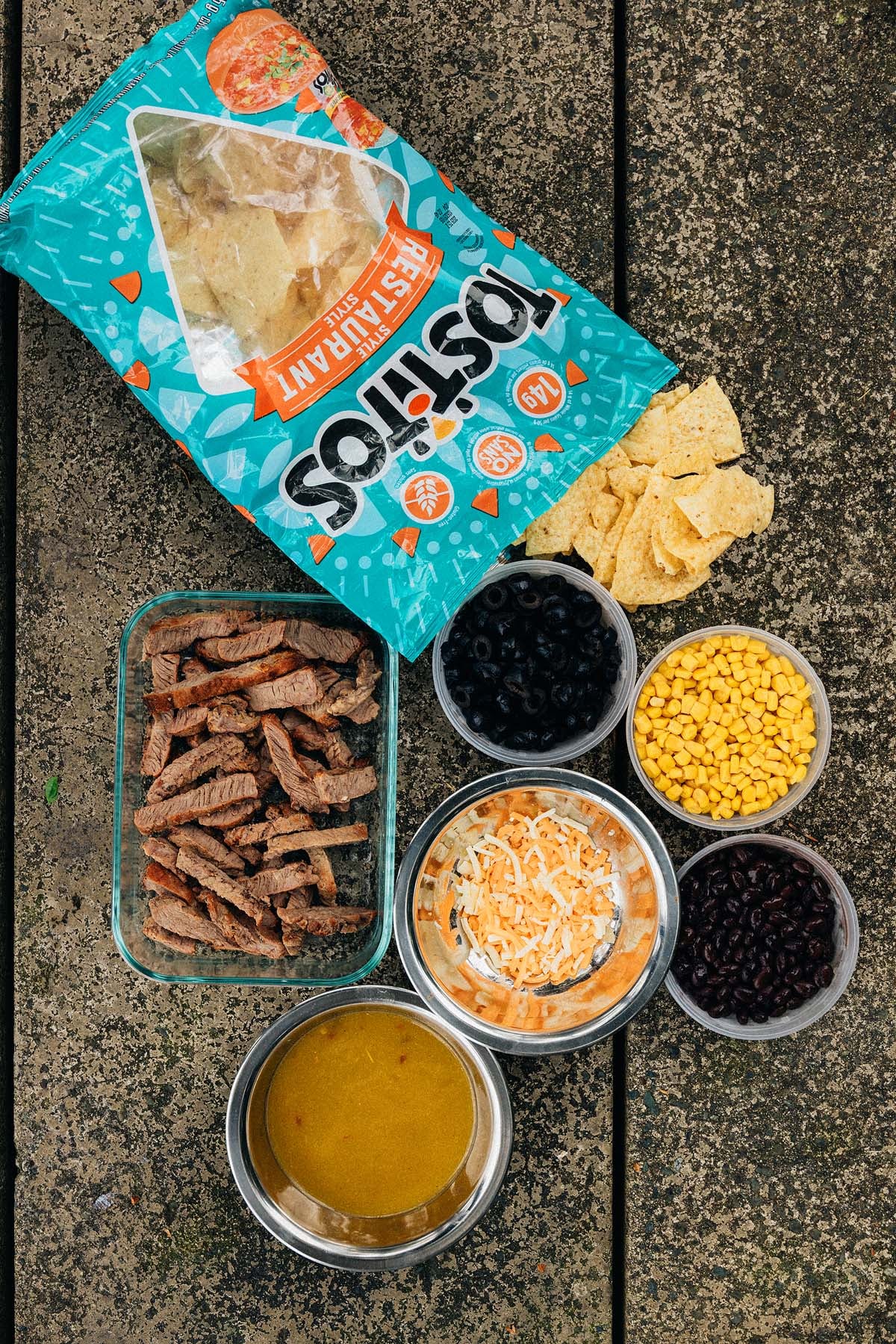 Open Tostitos chips bag with bowls of steak, shredded cheese, beans, corn, black olives, and sauce on a wooden surface&mdash;perfect for assembling Campfire Steak and Black Bean Nachos.
