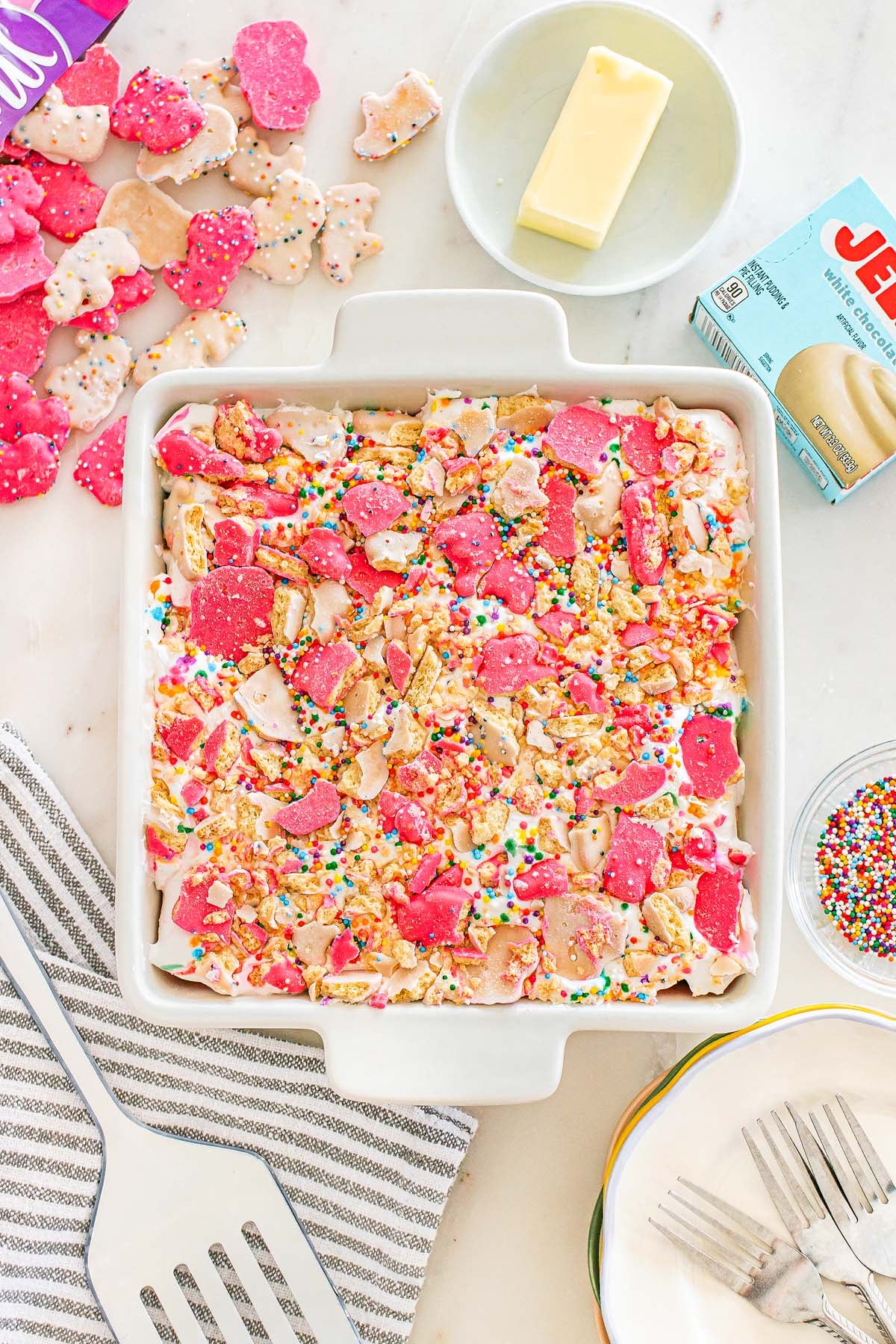 Square pan filled with Circus Animal Cookie Lush dessert, sprinkled with animal crackers and surrounded by ingredients and utensils on a white surface.