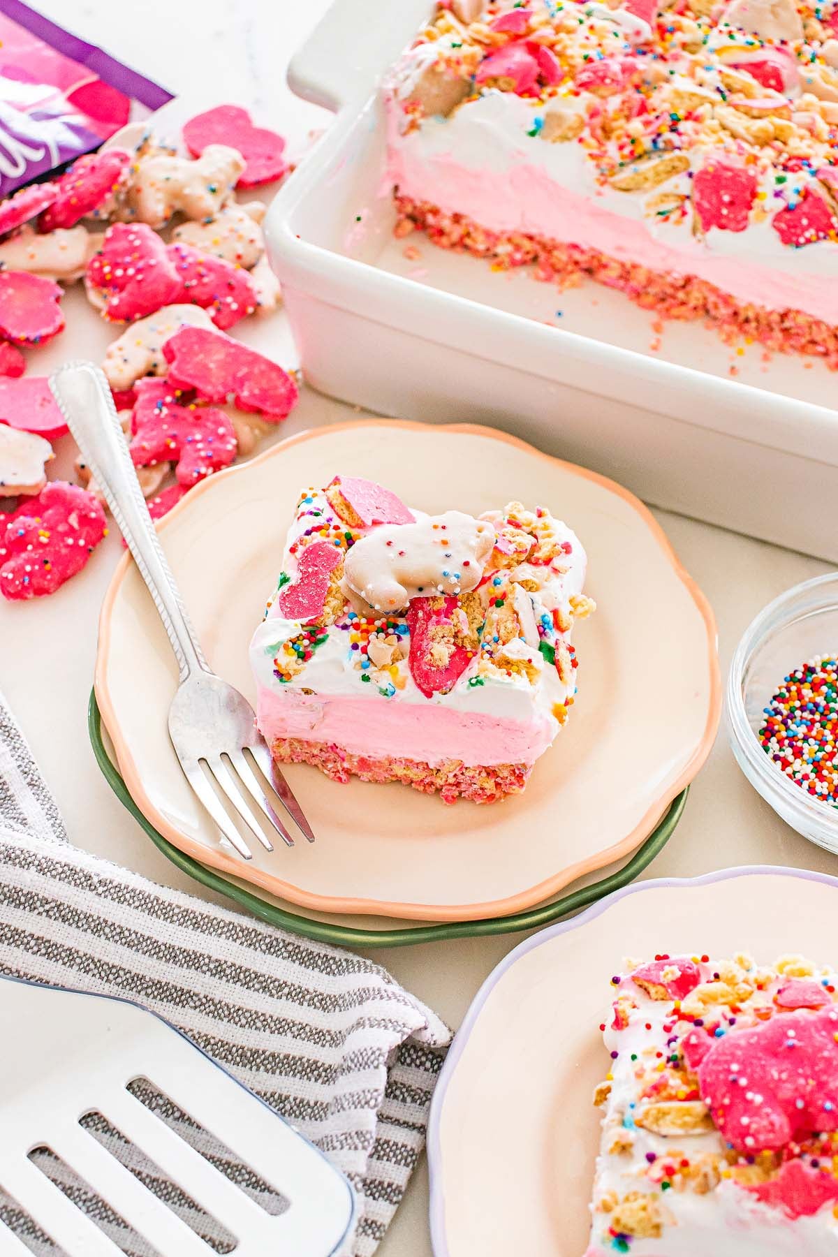 A pink and white Circus Animal Cookie Lush dessert topped with frosted animal cookies and sprinkles, served on a plate with a fork.