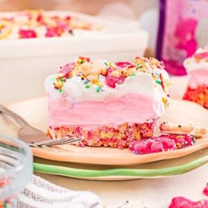 A slice of pink ice cream cake topped with sprinkles and frosted animal cookies, inspired by Circus Animal Cookie Lush, served on a plate with a fork.