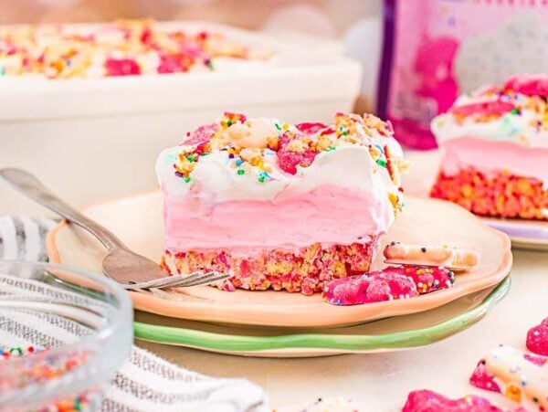 A slice of pink ice cream cake topped with sprinkles and frosted animal cookies, inspired by Circus Animal Cookie Lush, served on a plate with a fork.