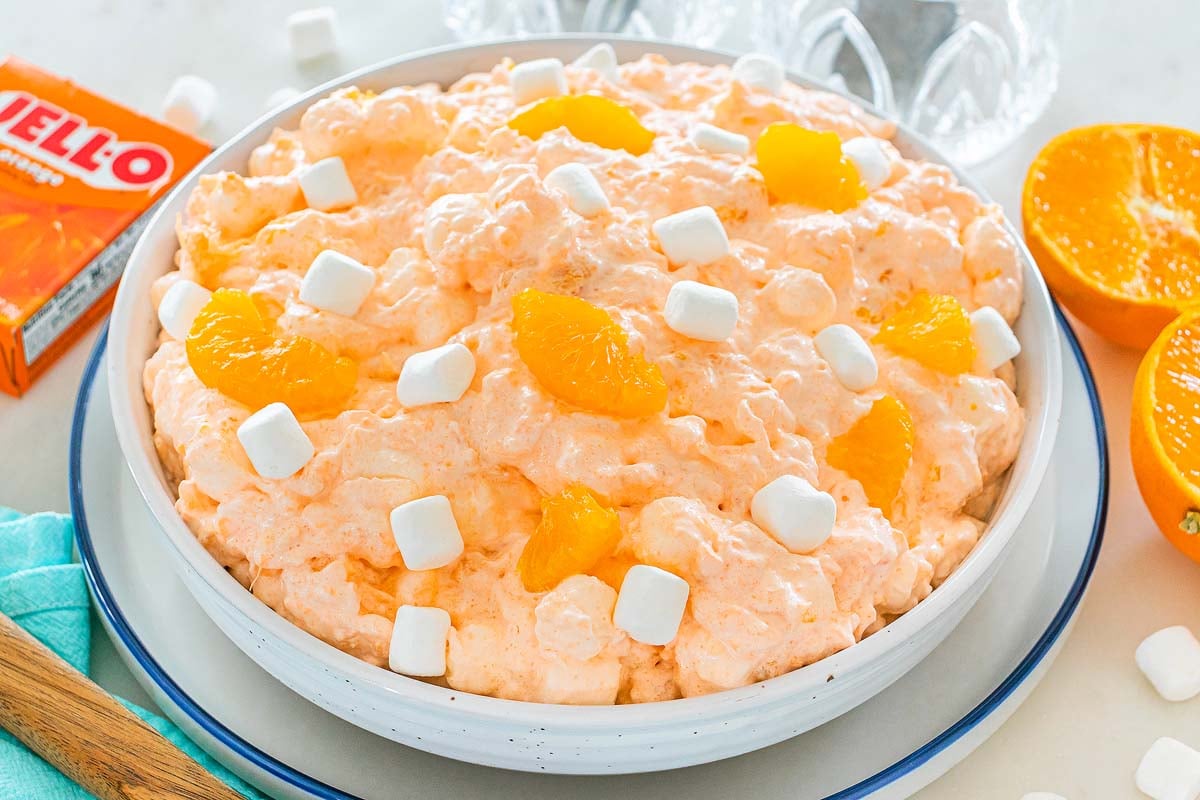 A bowl of Cottage Cheese Jello Salad, featuring orange fluff with mandarin slices and mini marshmallows on top, sits next to fresh oranges and Jell-O.