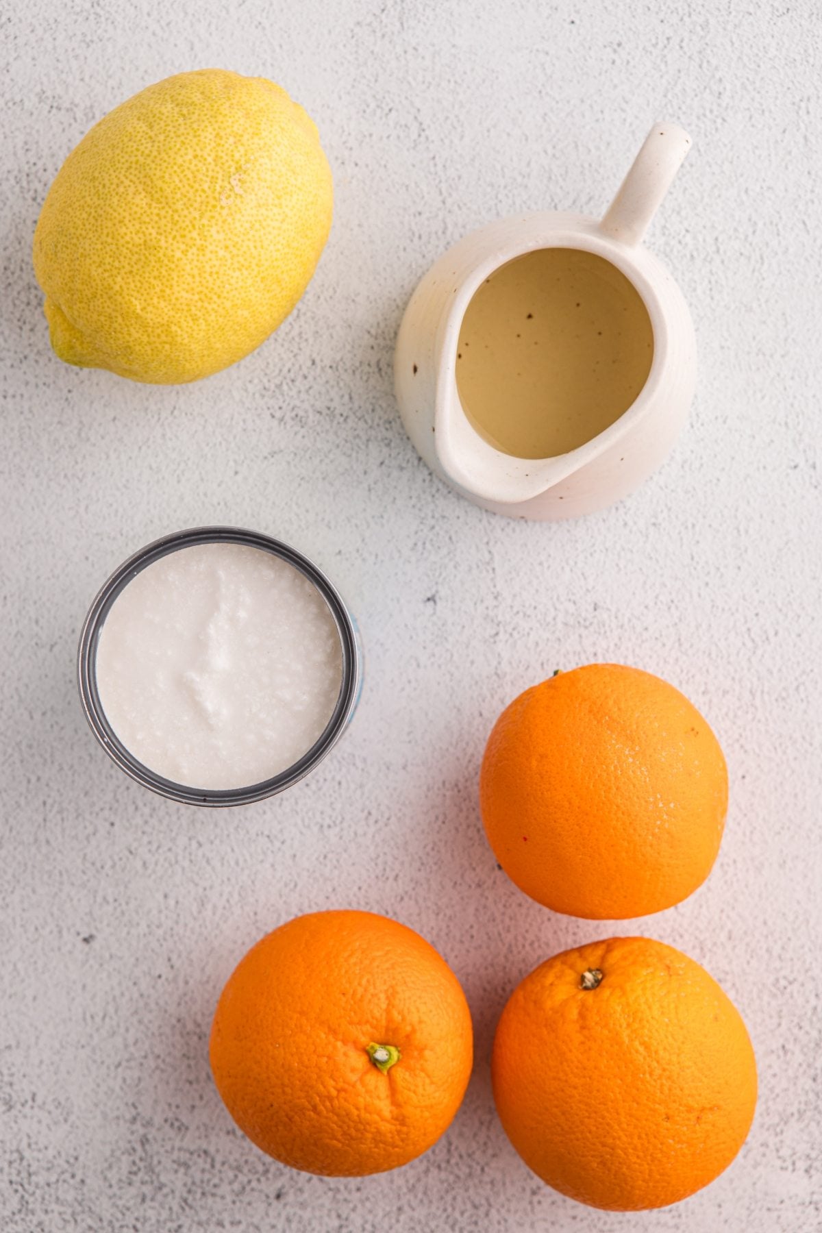 A lemon, three oranges, a small pitcher of liquid, and a jar of white cream on a light surface—perfect ingredients for refreshing orange sorbet.