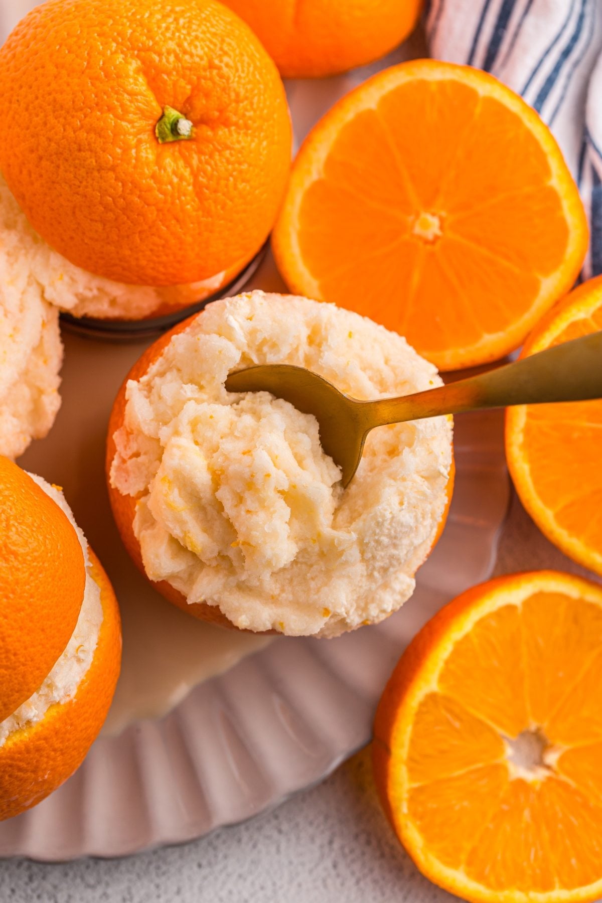 A gold spoon scooping creamy orange sorbet from a hollowed orange, surrounded by fresh orange halves.