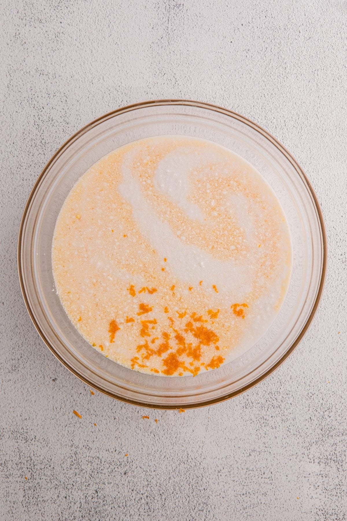 A glass bowl with milk, sugar, and orange zest—perfect ingredients for a refreshing orange sorbet—sits on a light gray textured surface.