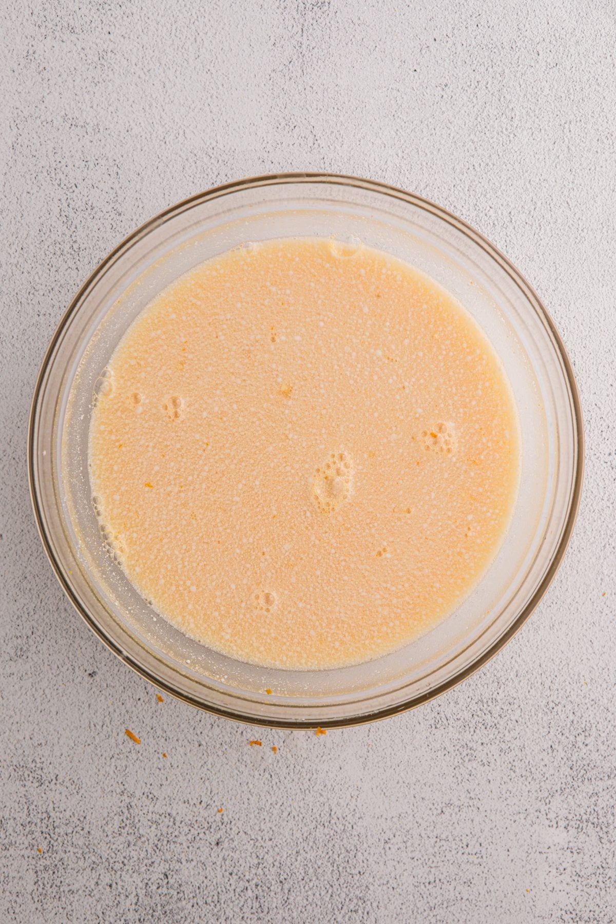 A glass bowl filled with a pale yellow, bubbly liquid mixture—reminiscent of freshly churned orange sorbet—sits atop a light gray surface.