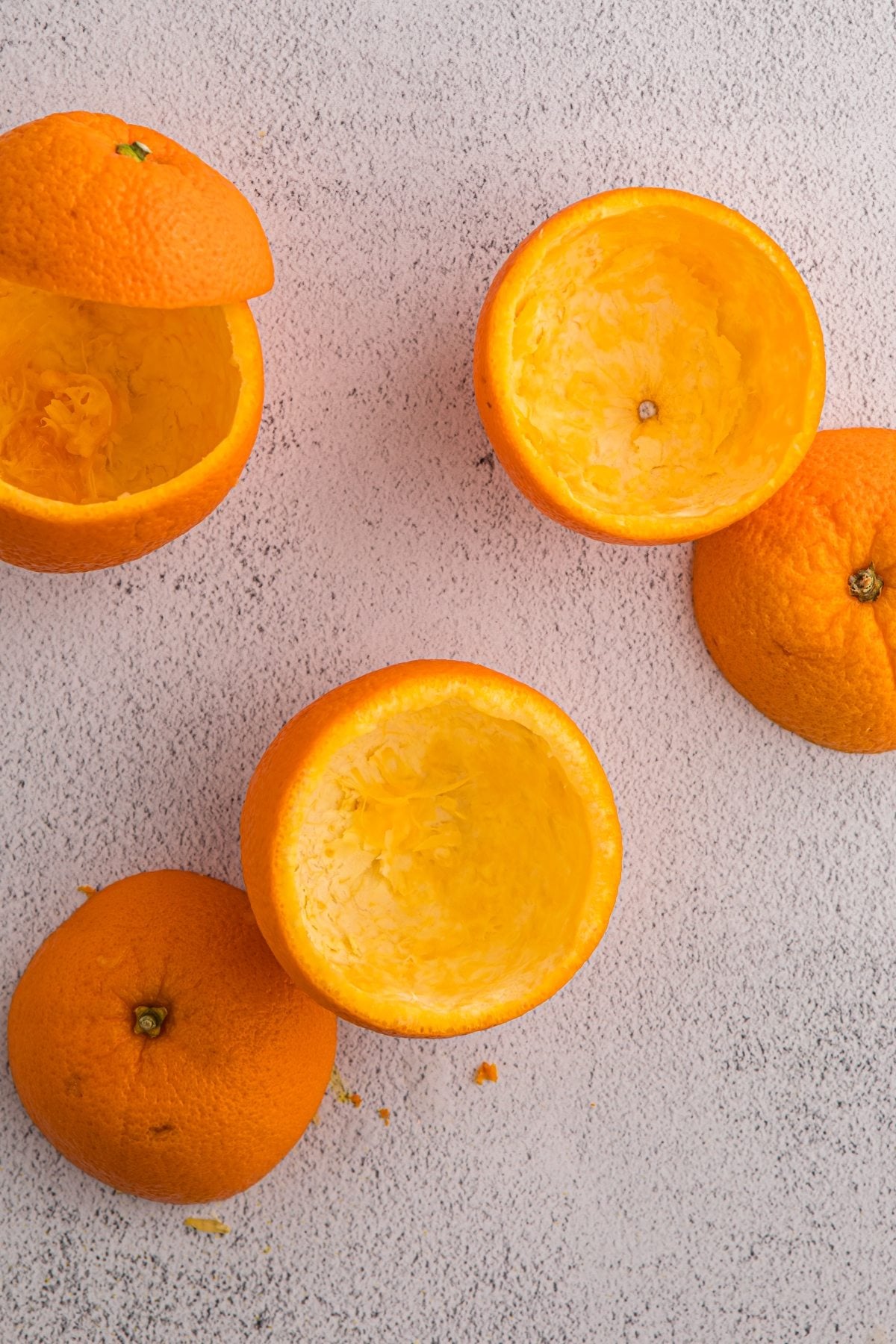 Four oranges, two whole and two hollowed out with their tops removed, ready to be filled with homemade orange sorbet, on a light textured surface.