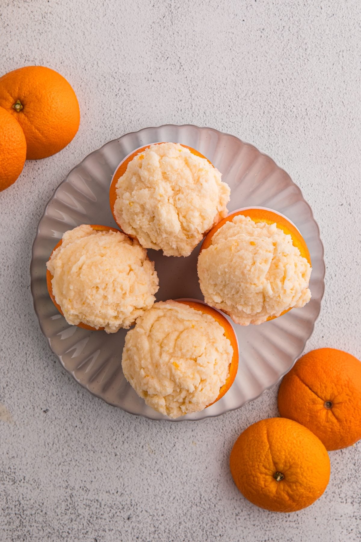 Four orange halves filled with creamy orange sorbet on a gray plate, surrounded by whole oranges.