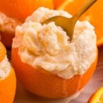Orange halves filled with creamy orange sorbet, one with a gold spoon, surrounded by more oranges.