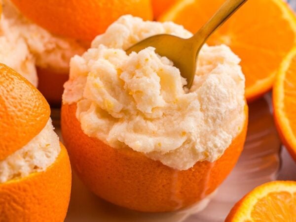 Orange halves filled with creamy orange sorbet, one with a gold spoon, surrounded by more oranges.
