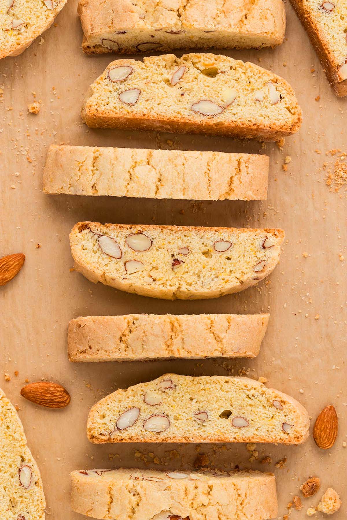 Sliced Almond Biscotti arranged in a row on parchment paper, with a few loose almonds nearby.