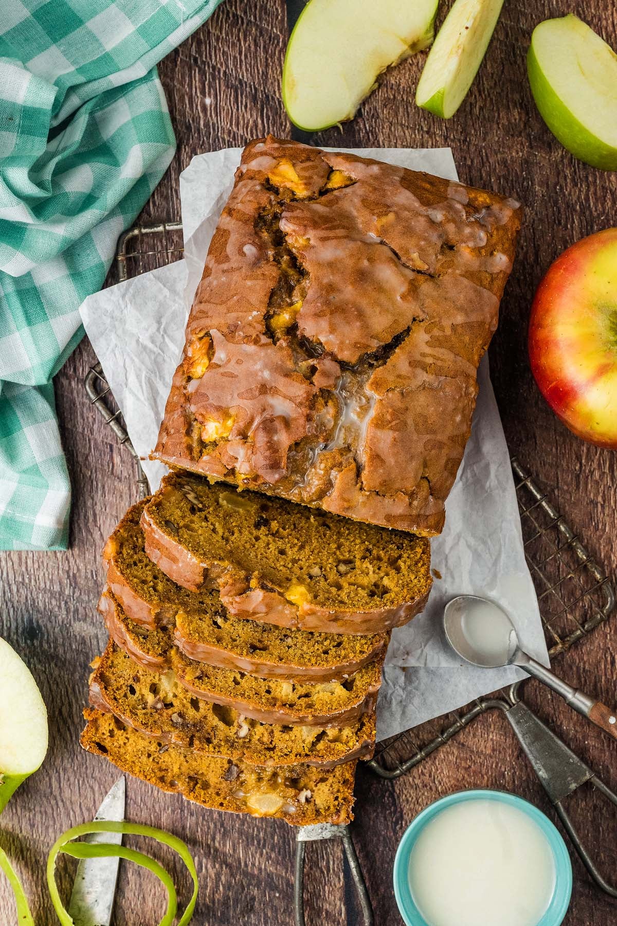 A sliced loaf of apple pumpkin bread on parchment paper, surrounded by apple slices and a green checkered cloth.