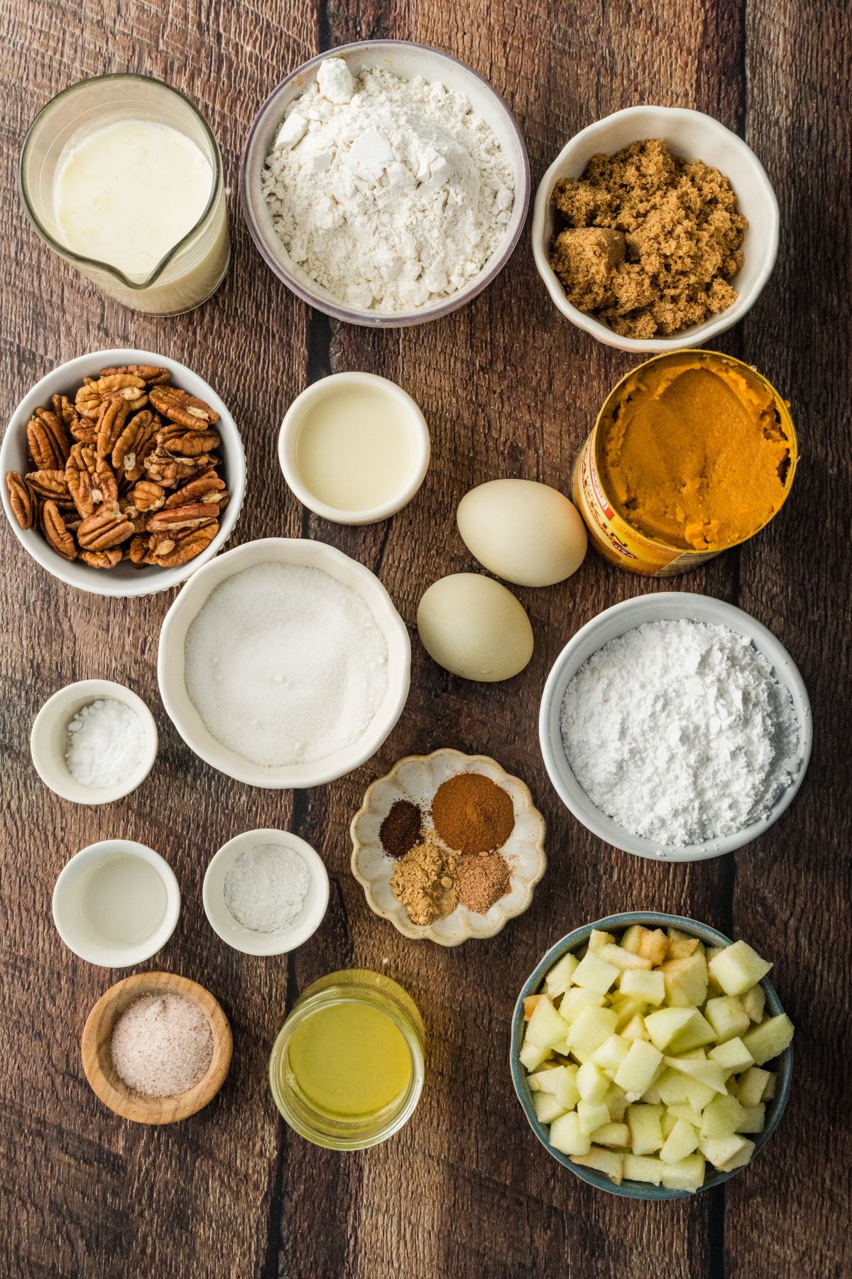 Assorted baking ingredients—flour, sugar, eggs, pumpkin puree, pecans, and spices—arranged on a wooden surface.