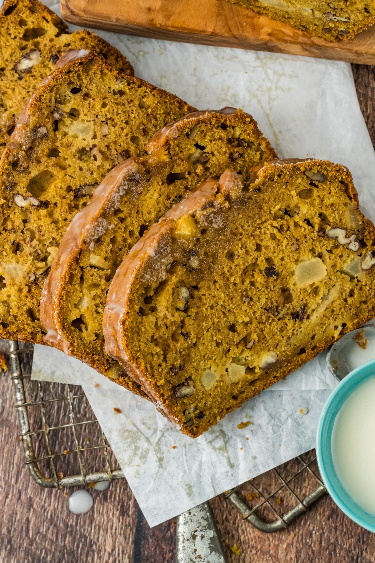 Slices of glazed apple bread with nuts on parchment paper, next to a small cup of white glaze.