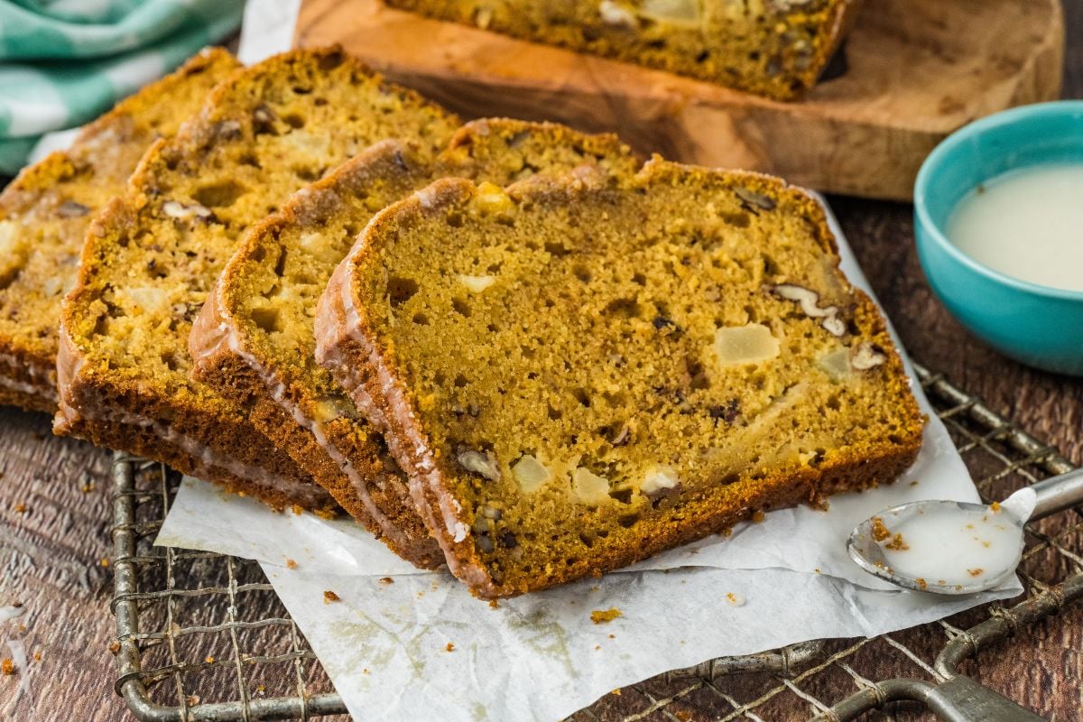 Slices of apple pumpkin bread with nuts and apple chunks rest on parchment paper, with a blue bowl in the background.