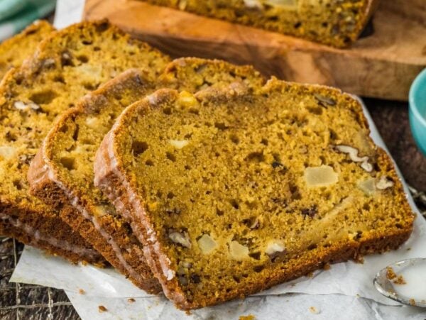 Slices of apple pumpkin bread with nuts and apple chunks rest on parchment paper, with a blue bowl in the background.