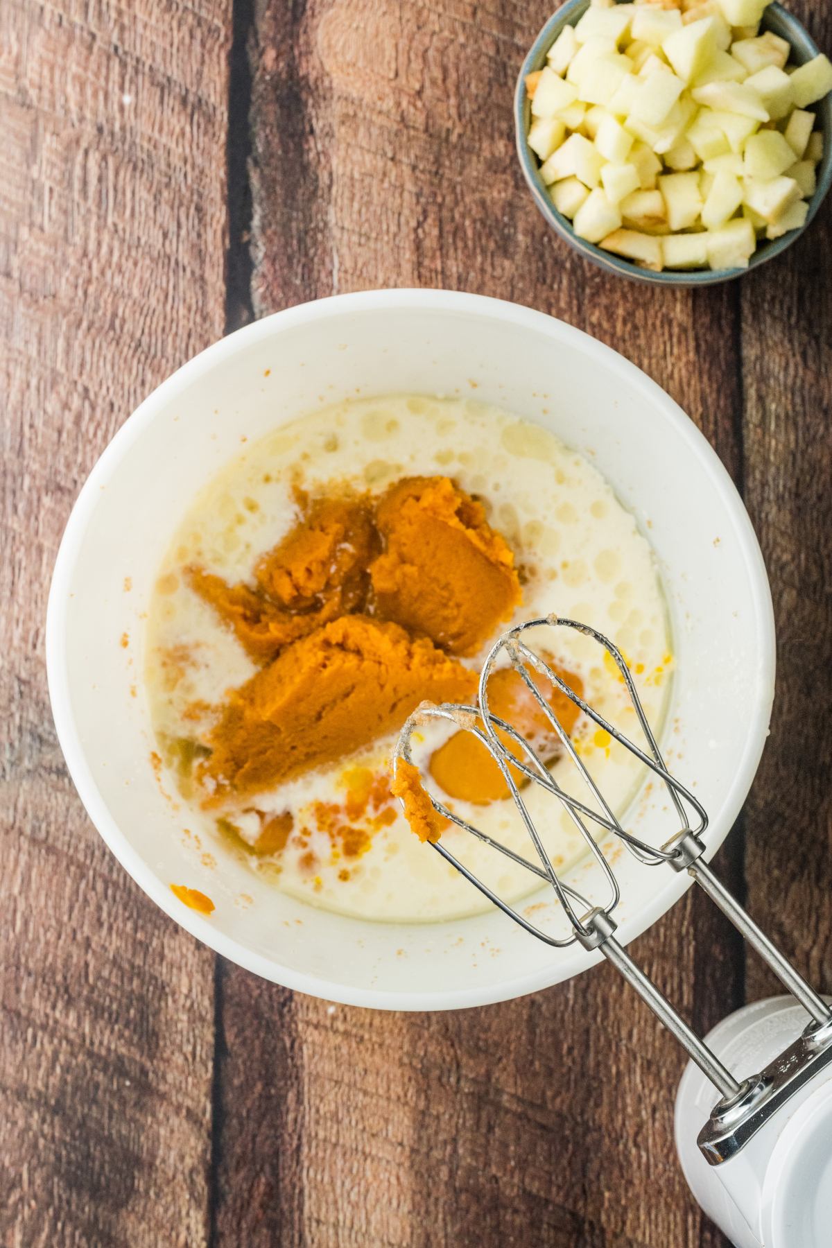 A mixing bowl with pumpkin, milk, and spices next to a hand mixer and a bowl of chopped apples.