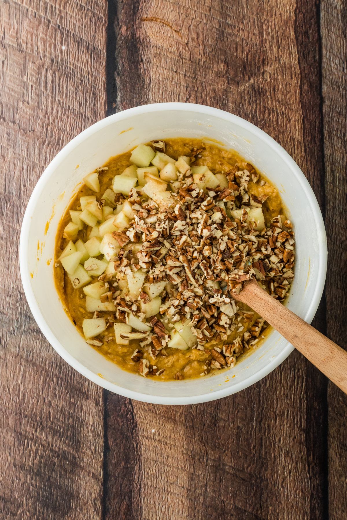 White bowl with chopped apples and pecans in batter, stirred with a wooden spoon on a wooden surface.