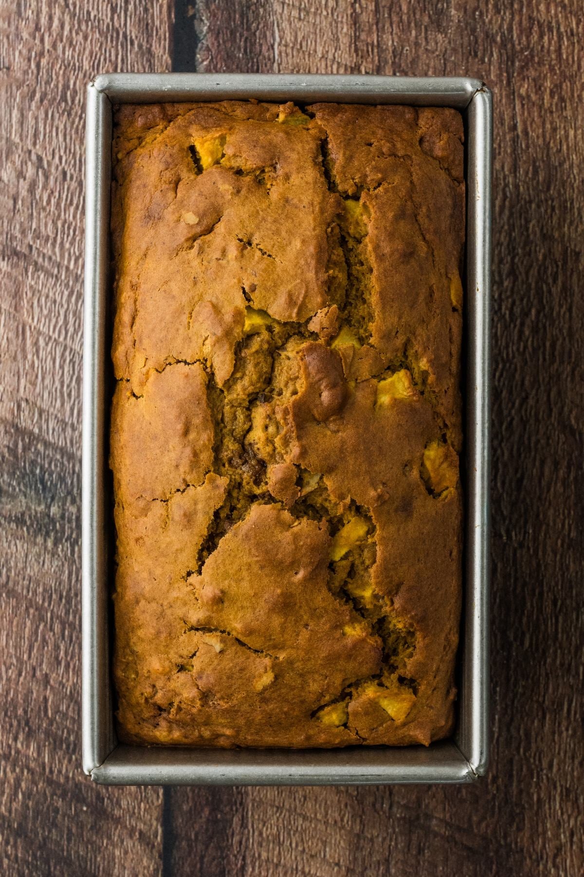 A loaf of pumpkin bread with a cracked top in a rectangular metal baking pan on a wooden surface.