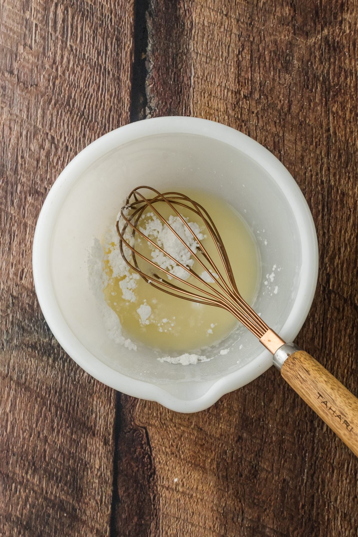 A white bowl with a whisk mixing powdered sugar and liquid on a wooden surface.