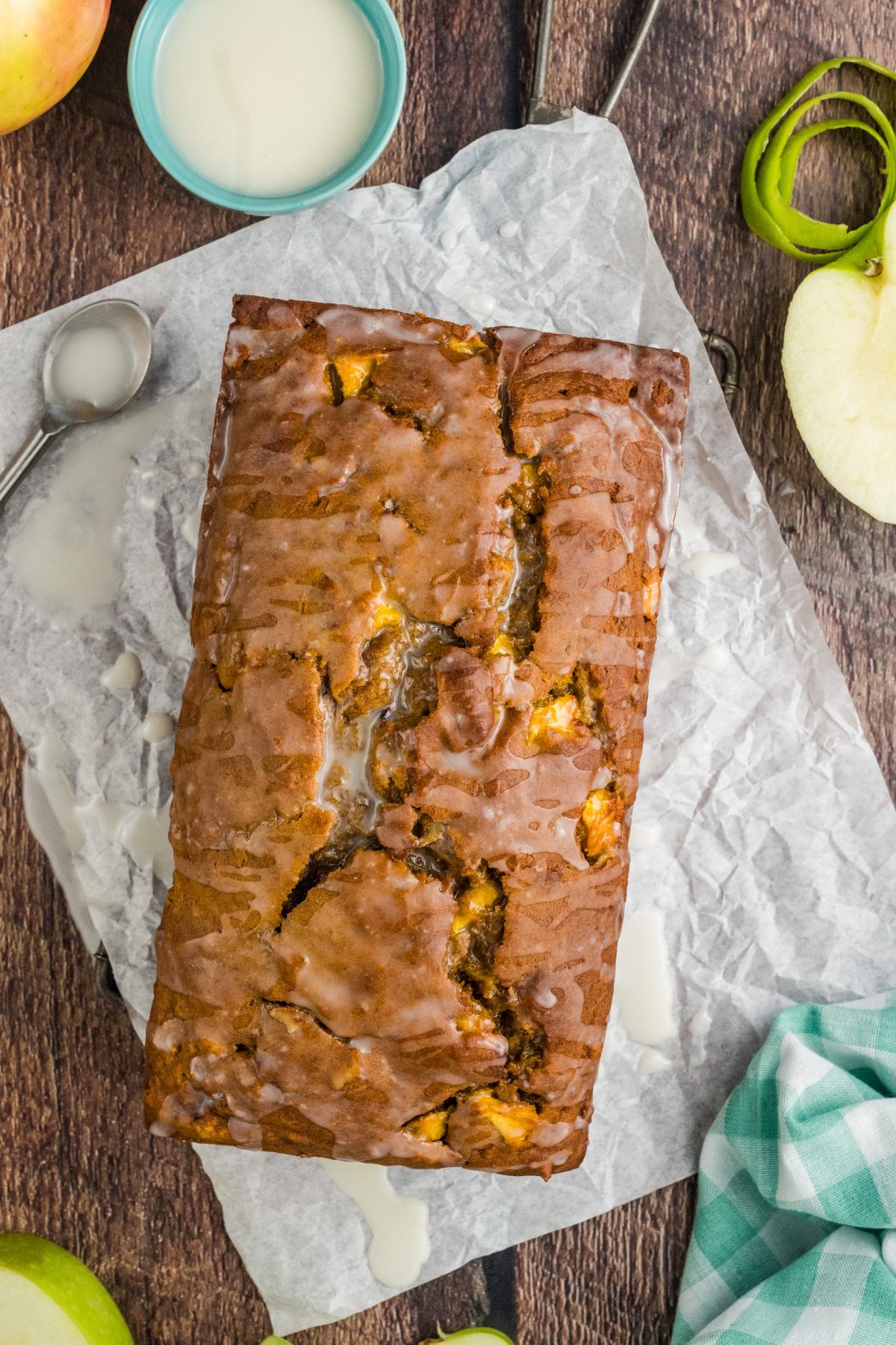 Glazed apple bread loaf on parchment paper, surrounded by apple slices and a blue bowl of icing.