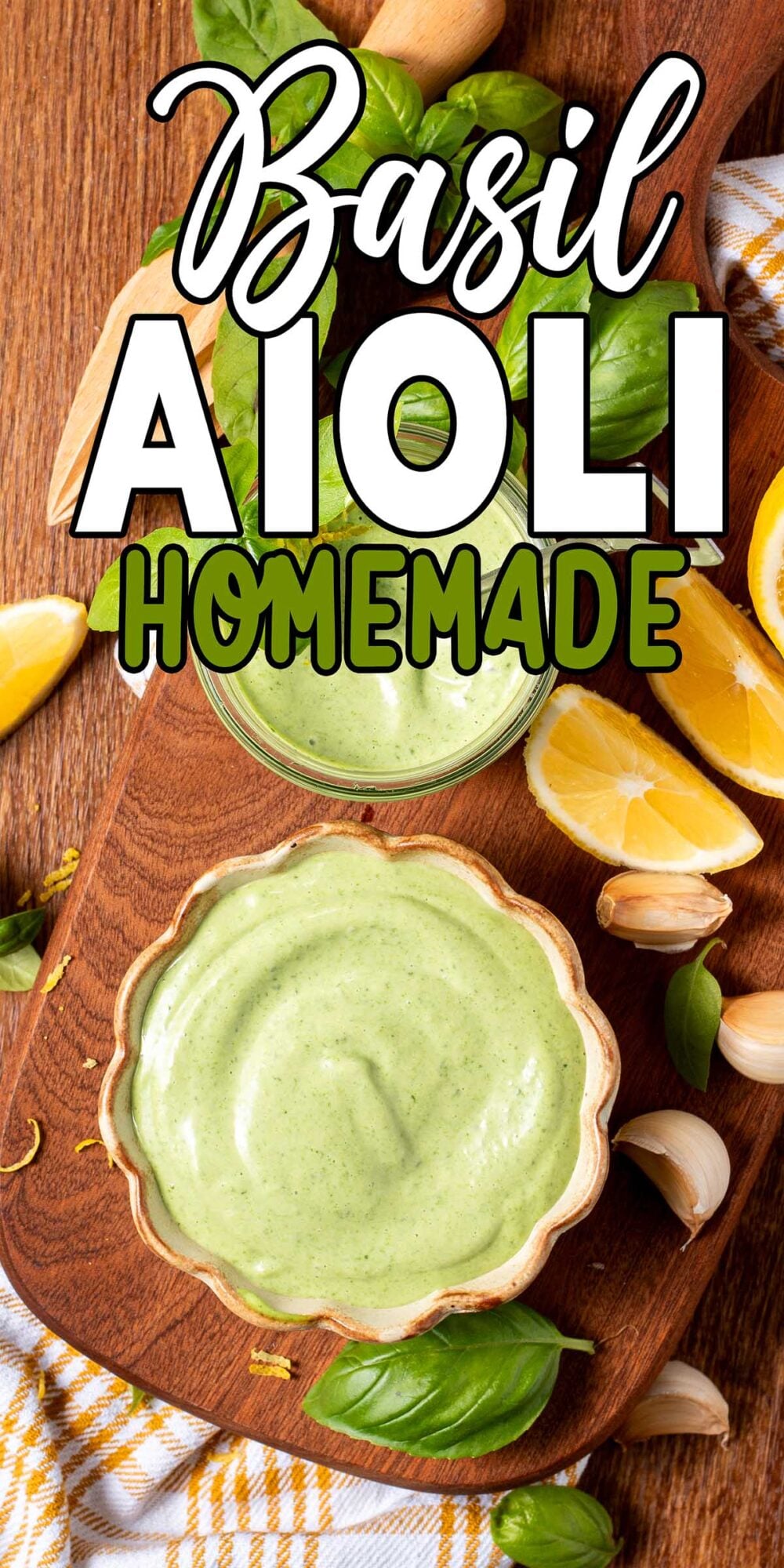 A bowl of creamy basil aioli on a wooden board, surrounded by lemon wedges, garlic cloves, and fresh basil leaves.