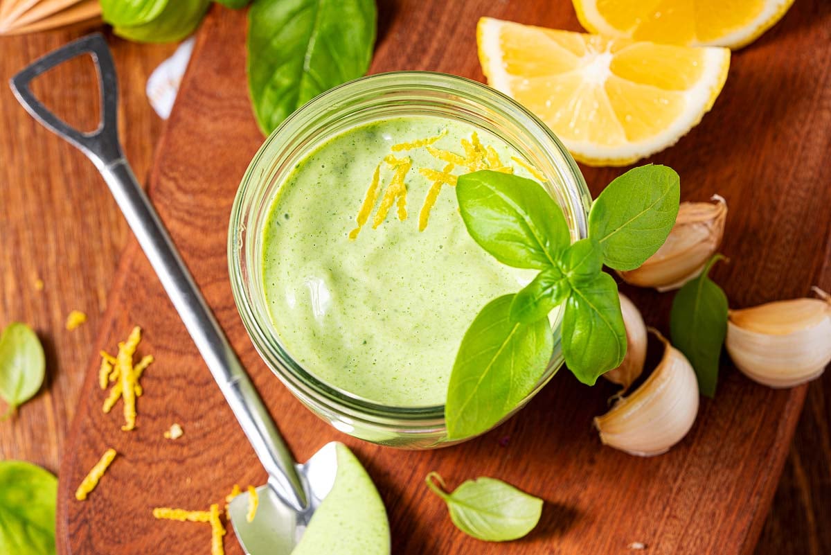 A jar of basil aioli garnished with lemon zest and basil, surrounded by lemon wedges, garlic, and fresh basil leaves.