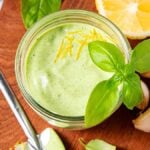 A jar of basil aioli garnished with fresh basil and lemon zest, surrounded by garlic, lemon, and vibrant basil leaves.