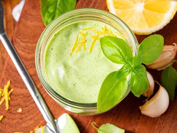 A jar of basil aioli garnished with fresh basil and lemon zest, surrounded by garlic, lemon, and vibrant basil leaves.