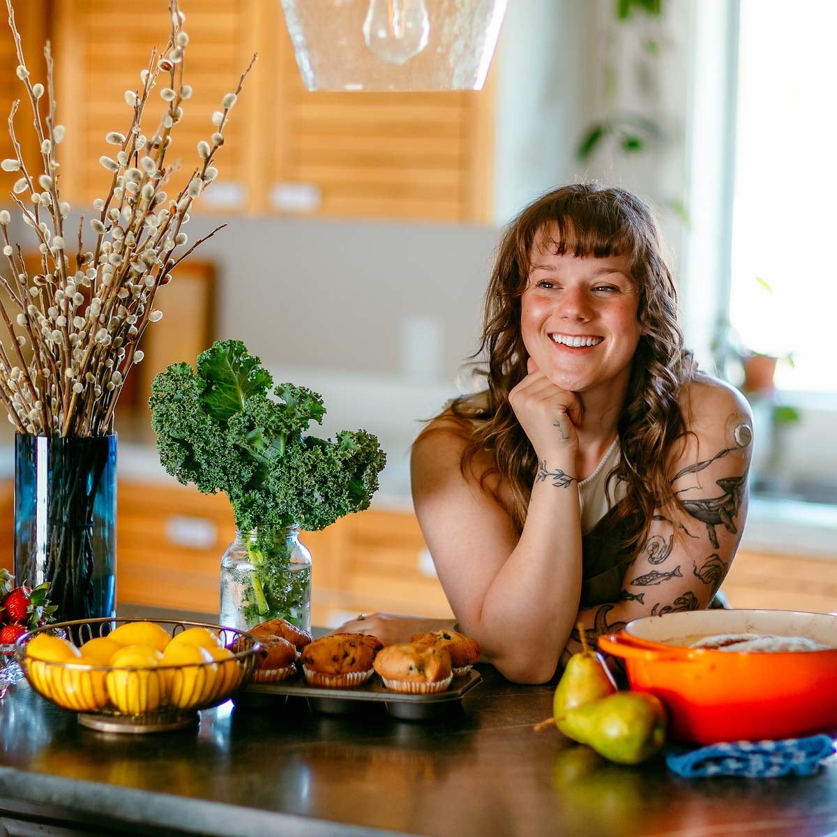 Woman smiling at a kitchen counter with fresh fruit, baked goods, kale, and a vase of branches.