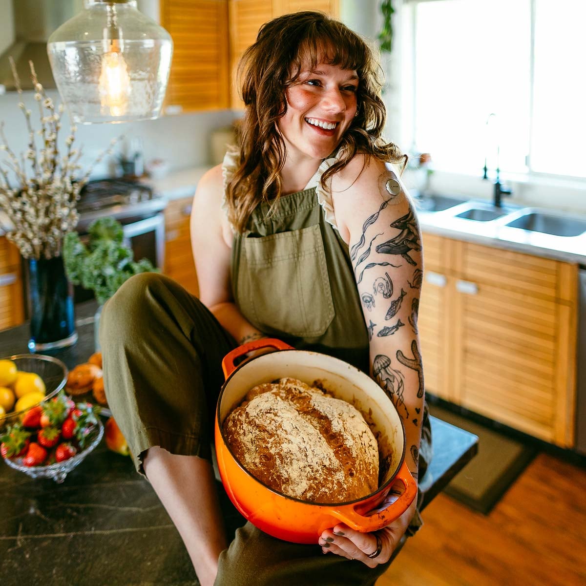 Smiling woman in an apron holds a pot with freshly baked bread in a bright kitchen.