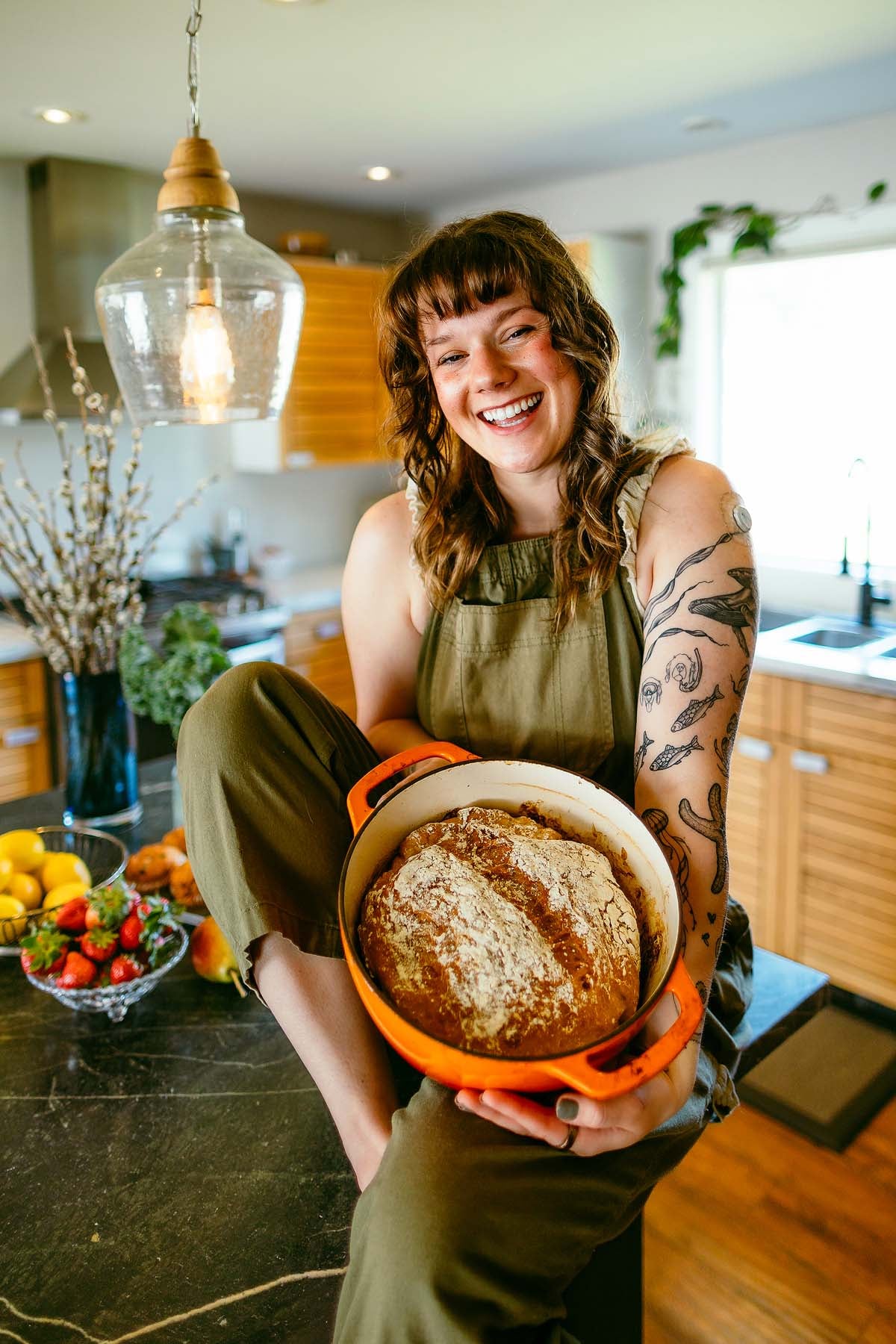 Smiling person in green overalls holding a pot of freshly baked bread in a bright kitchen.