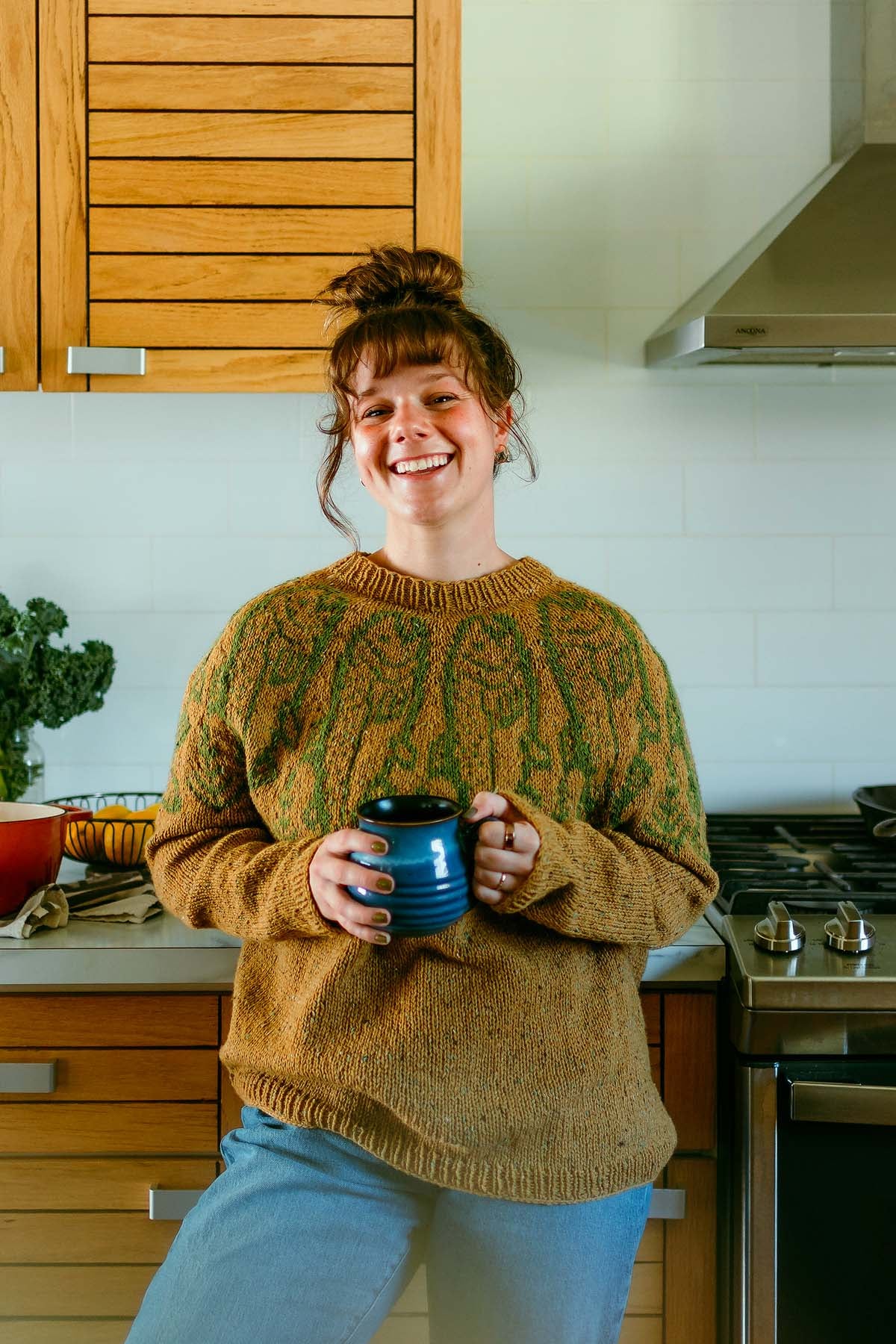 Bella Bucchiotti, in a cozy sweater, smiles while holding a blue mug in a modern kitchen with wooden cabinets.