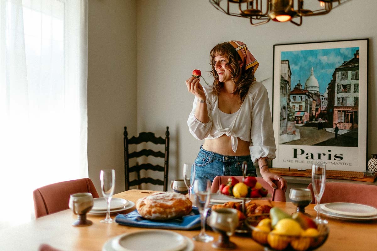 Bella Bucchiotti smiles in her kitchen, holding a strawberry by a table set with fresh fruit and pastries.