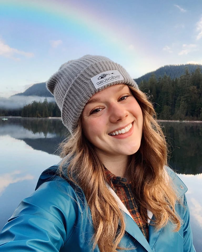 Smiling woman Bella Bucchiotti in a gray beanie stands by a lake, with a rainbow and forested mountains in the background.