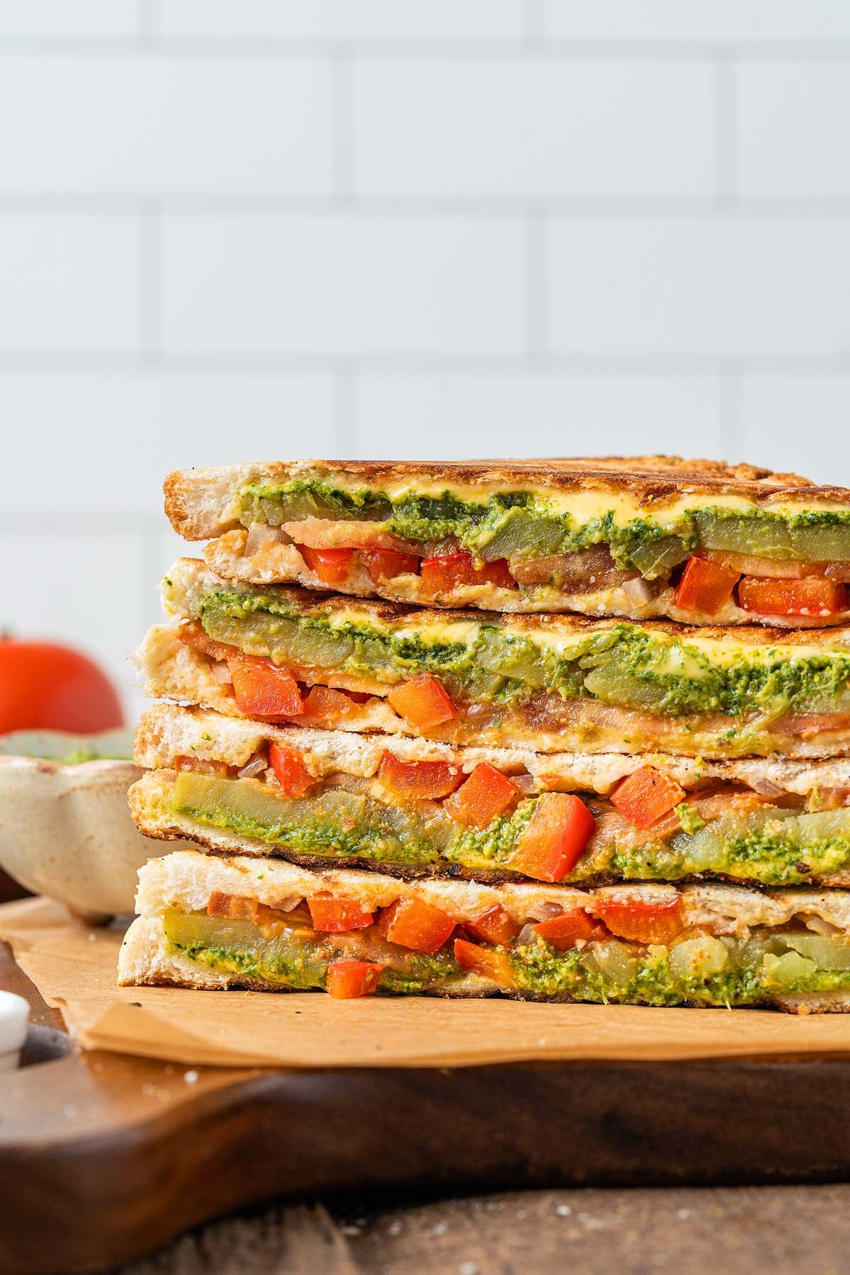 Stacked vegetable paninis with pesto, red peppers, and other veggies on a wooden board, inspired by classic Bombay sandwiches.