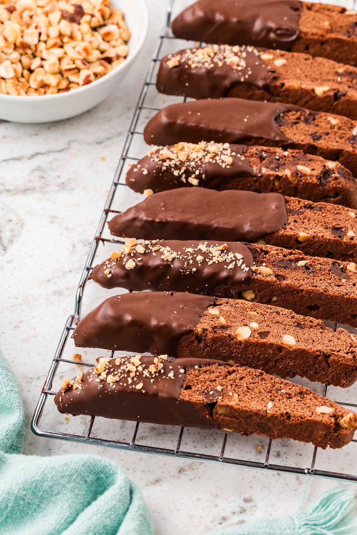Chocolate biscotti with nuts, dipped in rich chocolate, cools on a wire rack beside a bowl of chopped hazelnuts.