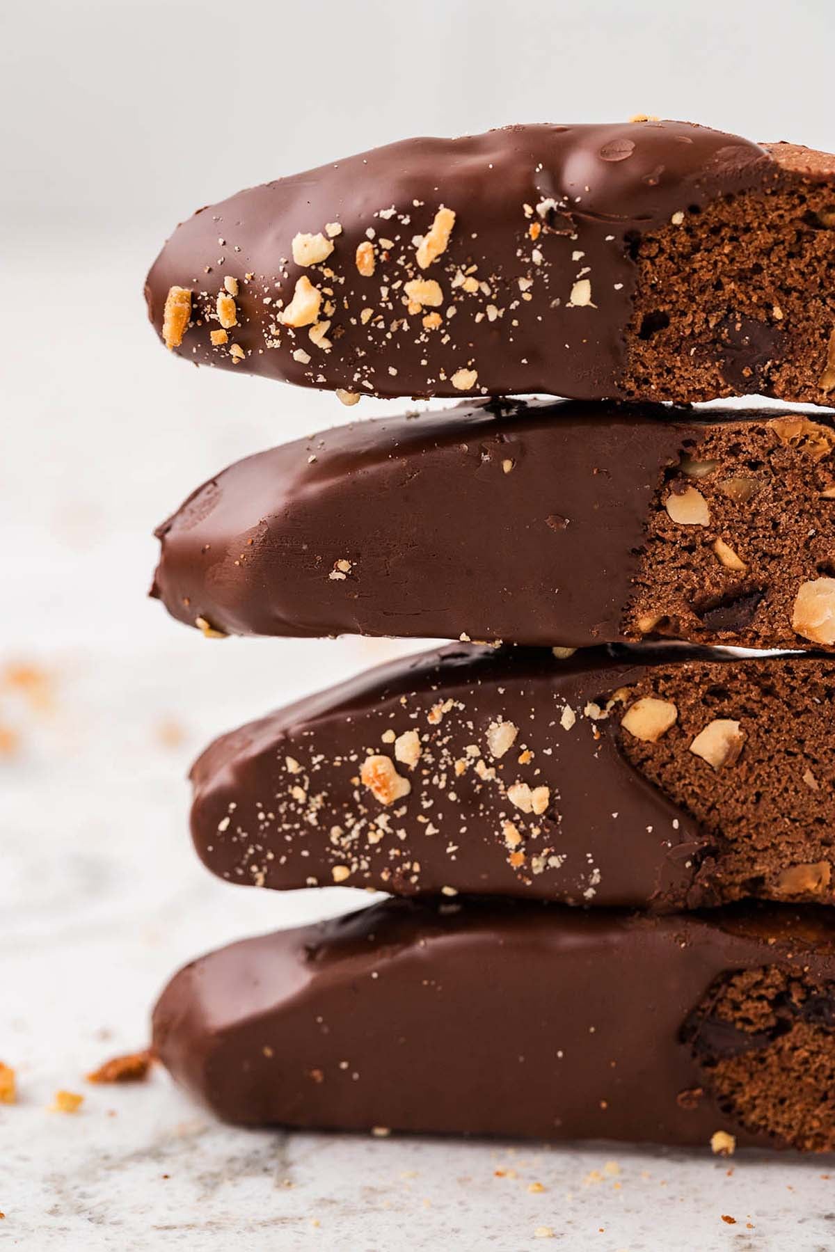 Four chocolate biscotti with chopped nuts are stacked on a light, speckled surface, each dipped in rich chocolate for a delicious treat.
