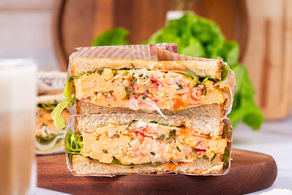 Close-up of a stacked Crab Sandwich with lettuce and creamy filling on a wooden board, wrapped in paper.