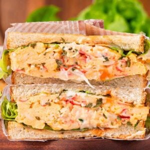 Two thick egg salad and crab sandwiches with lettuce, stacked on a wooden board, next to a glass of coffee.