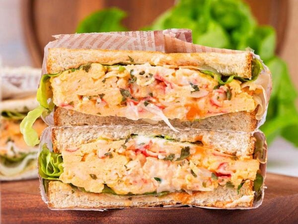 Two thick egg salad and crab sandwiches with lettuce, stacked on a wooden board, next to a glass of coffee.