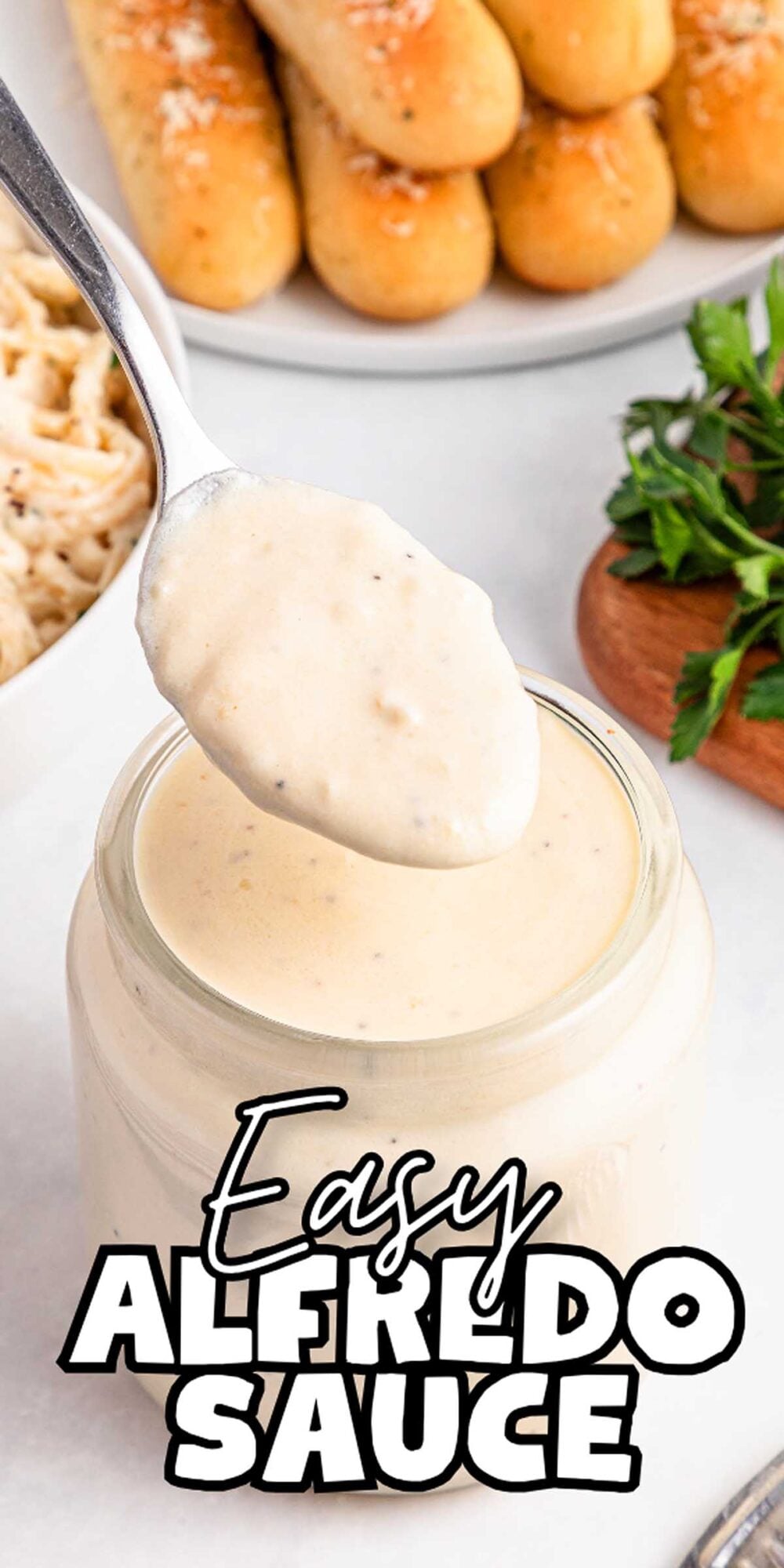 A spoonful of easy Alfredo sauce over a jar, with breadsticks and herbs in the background.