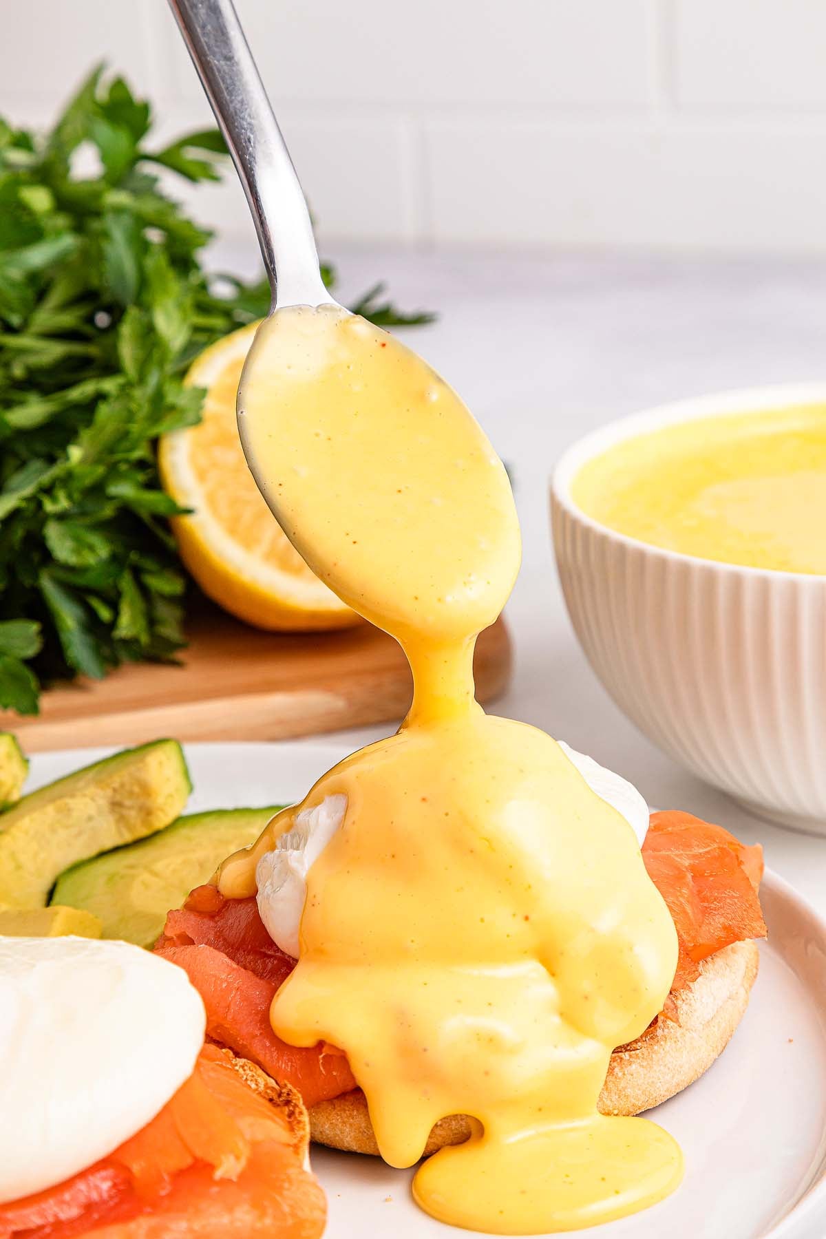 A spoon pours easy hollandaise sauce over eggs benedict with smoked salmon and avocado.